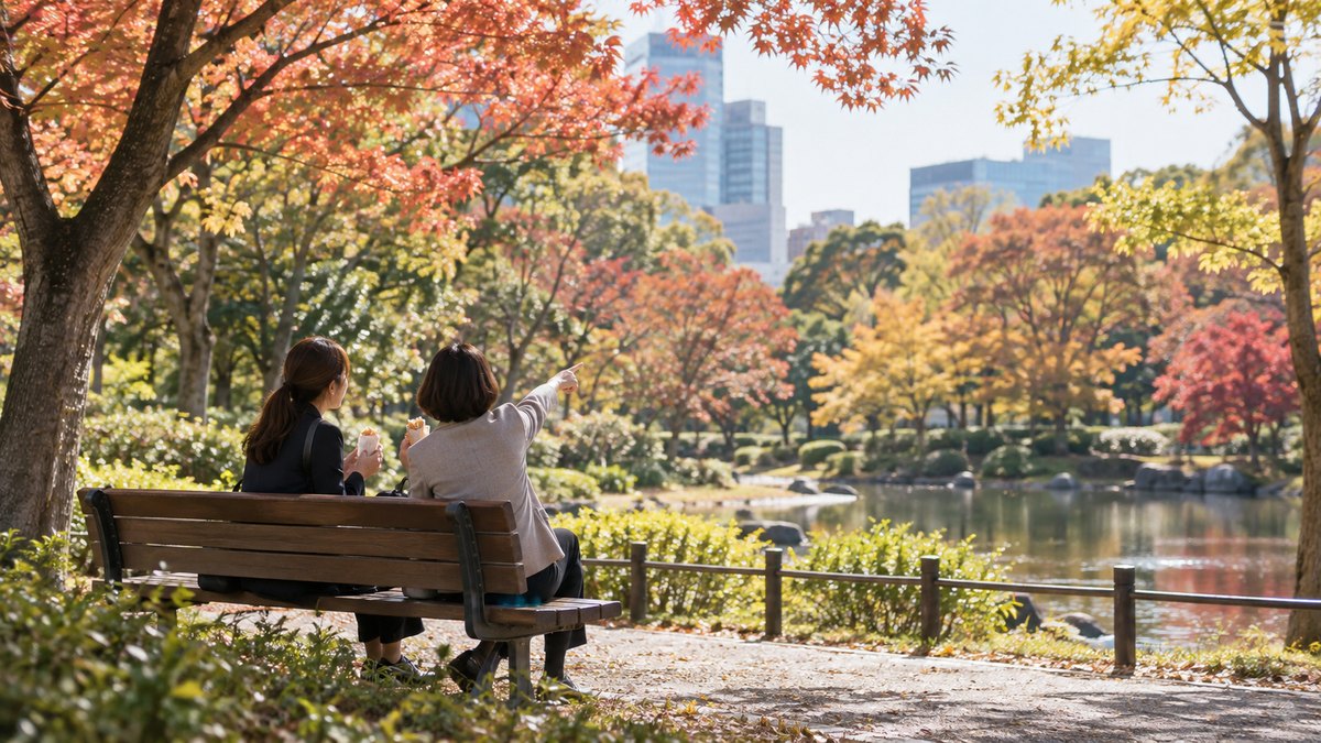 昼、会社の近くの公園のベンチで、30代の女性会社員と40代の女性の同僚が並んでサンドイッチを食べながら、色づいた木々を指さして話している遠景。顔ははっきり写らない。文字は無い