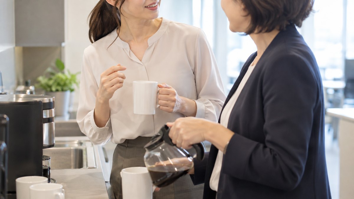 午前、オフィスの給湯室で、30代の女性会社員と40代の女性の同僚が並んでコーヒーを入れながら、女性が同僚のほうを向いて穏やかに一言話しかけている近景。顔ははっきり写らない。文字は無い