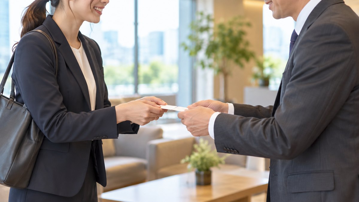 午後、取引先の会社の明るい応接スペースで、30代の女性会社員と40代の男性の担当者が向かい合って立ち、両手で名刺を交換しながら穏やかにほほえんでいる近景。顔ははっきり写らない。名刺や壁の文字は見えない
