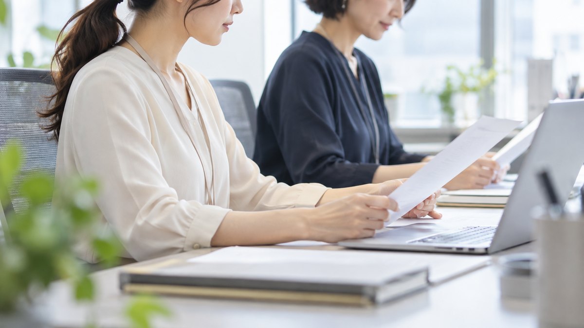 午前、明るいオフィスの共有デスクで、30代の女性会社員と40代の女性の同僚が隣り合って座り、言葉を交わさずにそれぞれ書類の確認を進めながら、落ち着いた穏やかな表情をしている横からの近景。顔ははっきり写らない。書類やパソコンの画面の文字は見えない