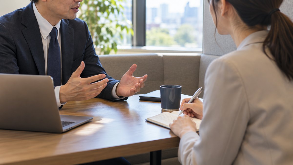 午後、オフィスの小さな打ち合わせブースで、40代の男性の上司と30代の女性の部下が向かい合って座り、部下が上司の話にメモを取りながらうなずいている近景。顔ははっきり写らない。メモと壁の文字は読めない