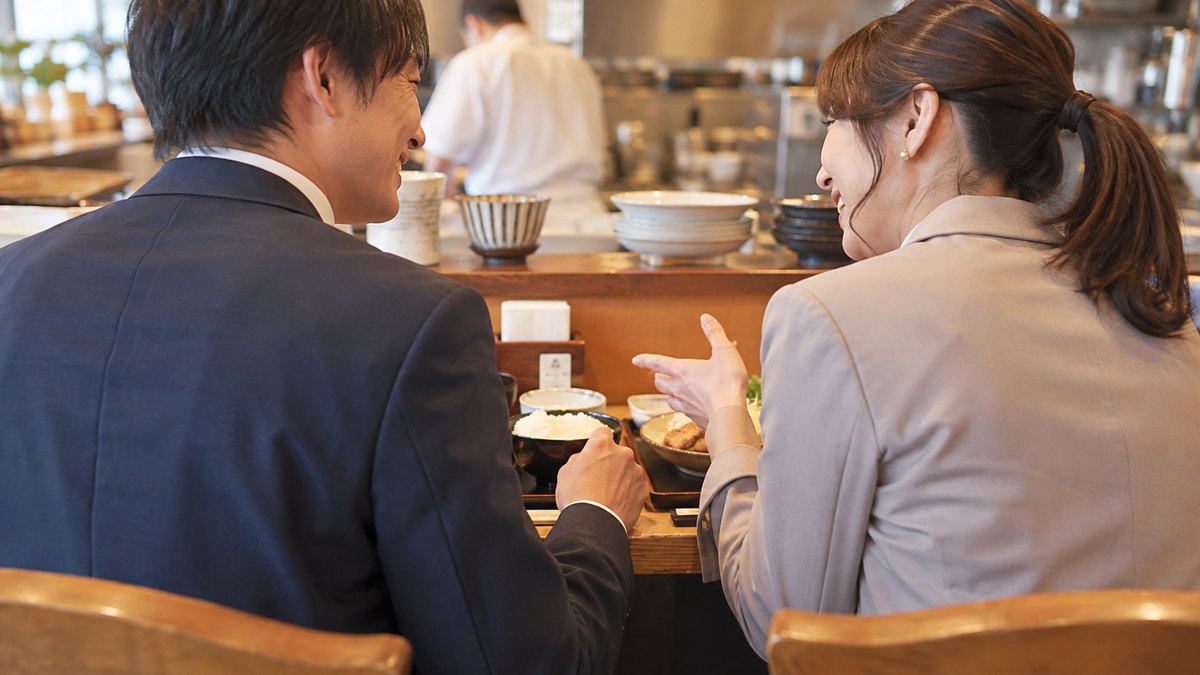 昼、会社の近くの小さな定食屋のカウンター席で、異動してきたばかりの30代の男性会社員が、隣に座った40代の女性の同僚におすすめの料理を教えてもらいながら笑顔で話を聞いている近景。顔ははっきり写らない。メニューや店の文字は見えない
