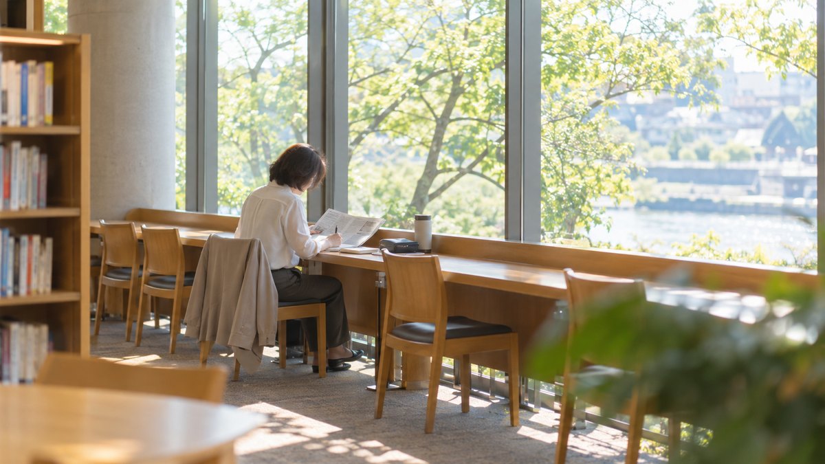 週末の朝、地域の図書館の窓ぎわの閲覧席で、50代の女性会社員が新聞のコラムを開き、ノートにゆっくり書き写している遠景。新聞とノートの文字は読めない。顔ははっきり写らない。文字は無い