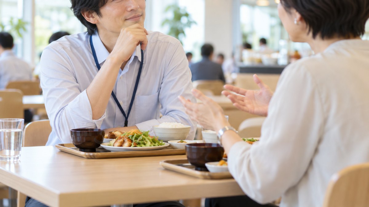 昼、社員食堂のテーブルで、30代の男性社員が定食を前に、向かいに座る40代の女性の先輩の話し方に耳を傾け、感心したようにうなずいている近景。メニューや掲示の文字は見えない。顔ははっきり写らない。文字は無い