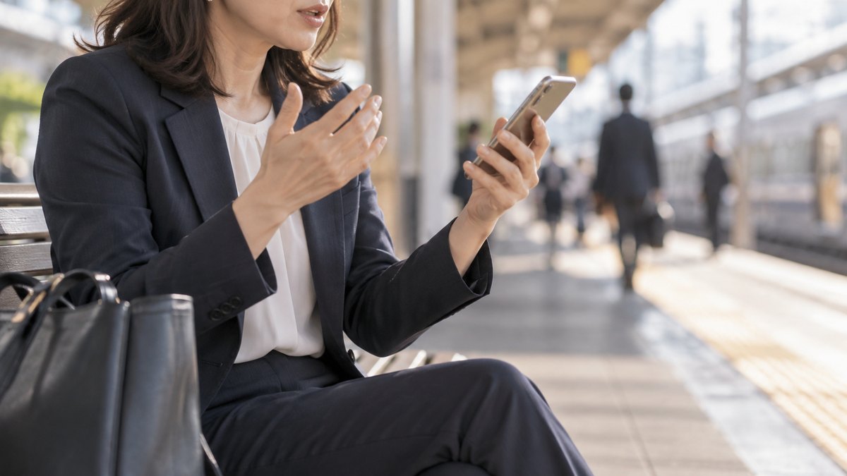 朝、駅のホームのベンチで、出勤前の40代の女性会社員がスマホのストップウォッチを手に、小さな声でスピーチの時間を測っている近景。スマホの画面は見えない。顔ははっきり写らない。文字は無い