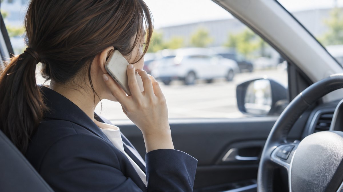 昼、会社の駐車場に停めた社用車の運転席で、30代の女性会社員がエンジンを止めたまま、スマホを耳に当てて自分の録音を聞き返している近景。スマホの画面は見えない。顔ははっきり写らない。文字は無い