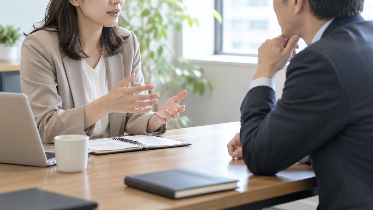 午後、明るい小さな会議室で、40代の男性の課長と30代の女性の部下がテーブルの角をはさんで斜めに座り、1対1で落ち着いて話している近景。部下が話し、課長がうなずいている。顔ははっきり写らない。ノートの文字は読めない