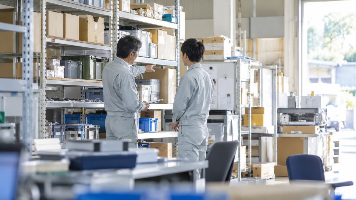 午前、明るい倉庫の事務スペースで、作業着の40代の男性の係長が、作業着の20代後半の男性の部下に棚の前で段取りを1つだけ説明している。部下はうなずきながら聞いている遠景。顔ははっきり写らない。棚や掲示の文字は読めない