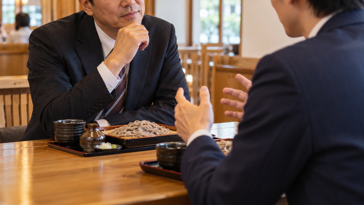 昼、会社の近くのそば屋のテーブル席で、40代の男性の課長と30代の男性の部下が向かい合って座り、課長が話を聞きながらうなずいている近景。顔ははっきり写らない。メニューや店の文字は見えない