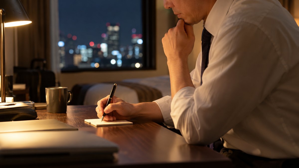 夜、出張先のビジネスホテルの部屋の机で、40代の男性会社員がスタンドの明かりの下、翌朝の会議で言う一言を小さなメモに書きながら口の中で確かめている横からの近景。顔ははっきり写らない。メモの文字は読めない