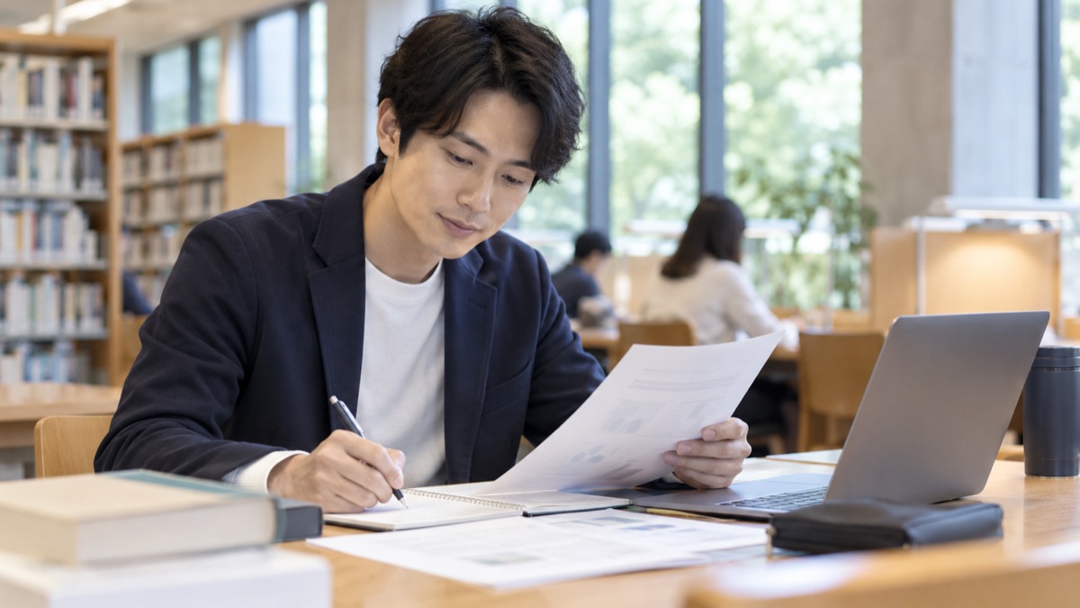 図書館の閲覧席で、会社の資料を読みながら質問をメモする20代後半の男性