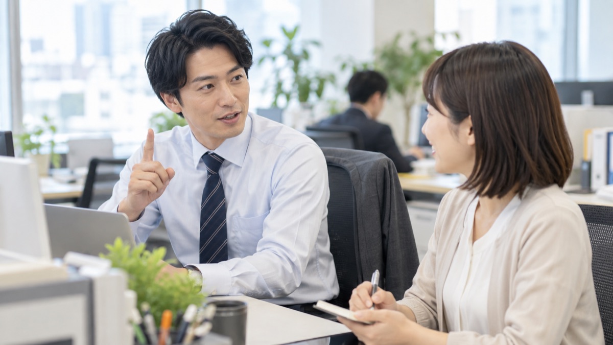 デスクで隣の同僚に指を1本立てて要点を短く伝える男性社員