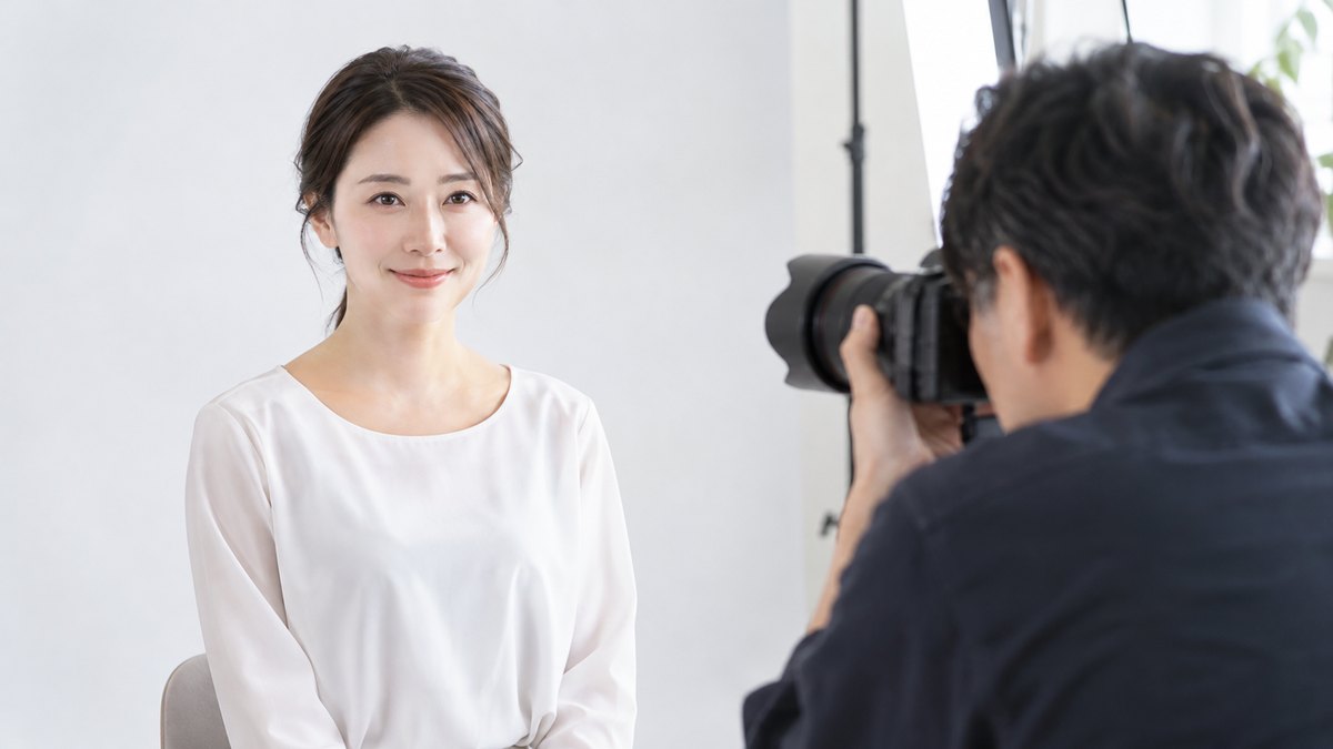 午後、明るい写真スタジオで、30代の女性が無地の背景の前に背筋を伸ばして座り、カメラマンの40代男性に向かって穏やかな表情で正面を向いている近景。カメラや背景に文字やロゴは無い