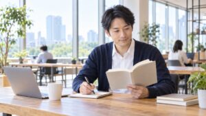 コワーキングスペースの机で、本を開いてノートに言葉を書き留める男性会社員