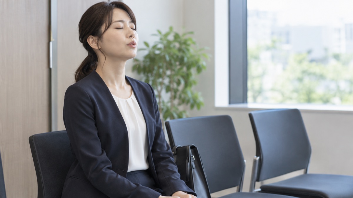 面接会場の控室の椅子に座り、目を閉じて口から細く長く息を吐く30代の女性