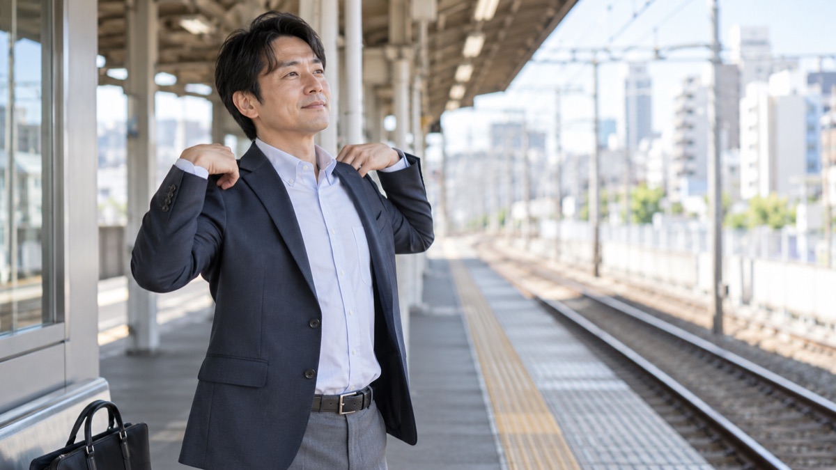 駅のホームの端で、電車を待ちながら肩をゆっくり回して体をほぐす40代の男性