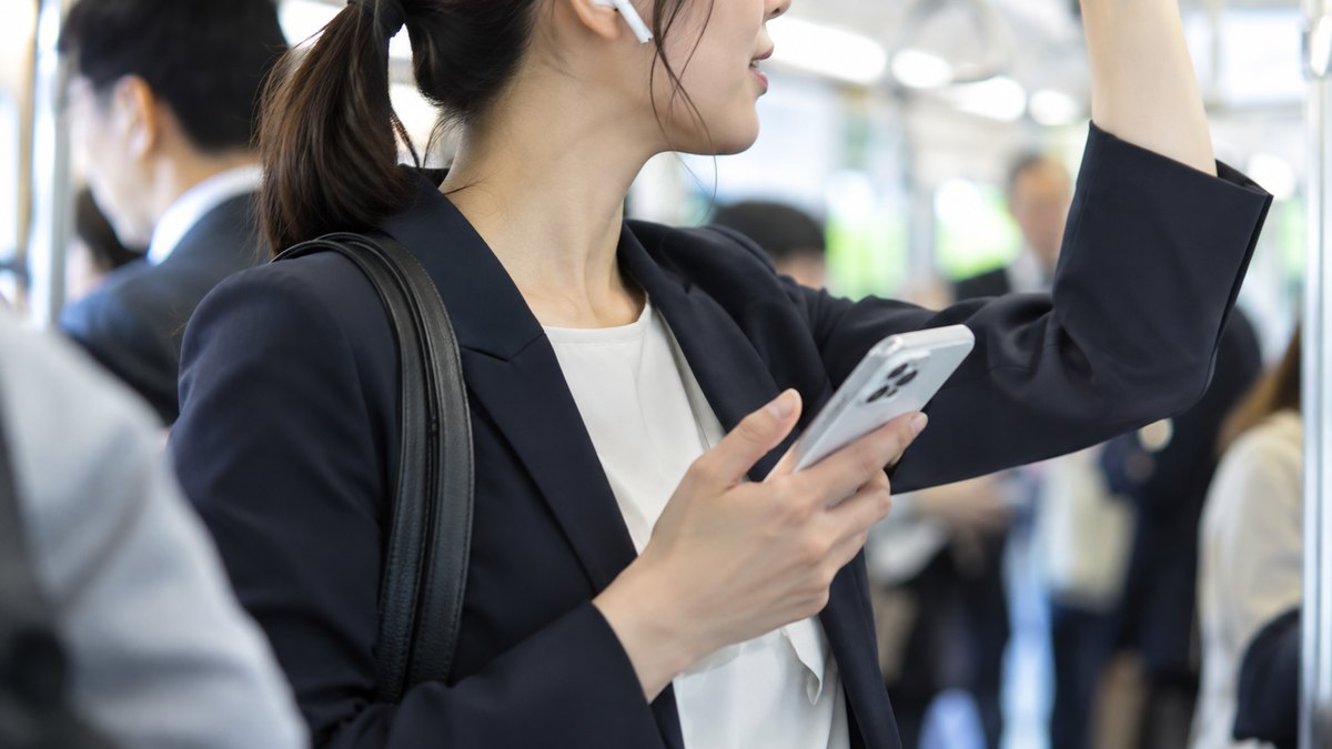 朝の通勤電車の中で、吊り革につかまった30代の女性会社員が片耳にイヤホンをつけ、スマホを胸の前で持って何かを聞き返している近景。顔ははっきり写らない。スマホの画面と車内の表示の文字は見えない
