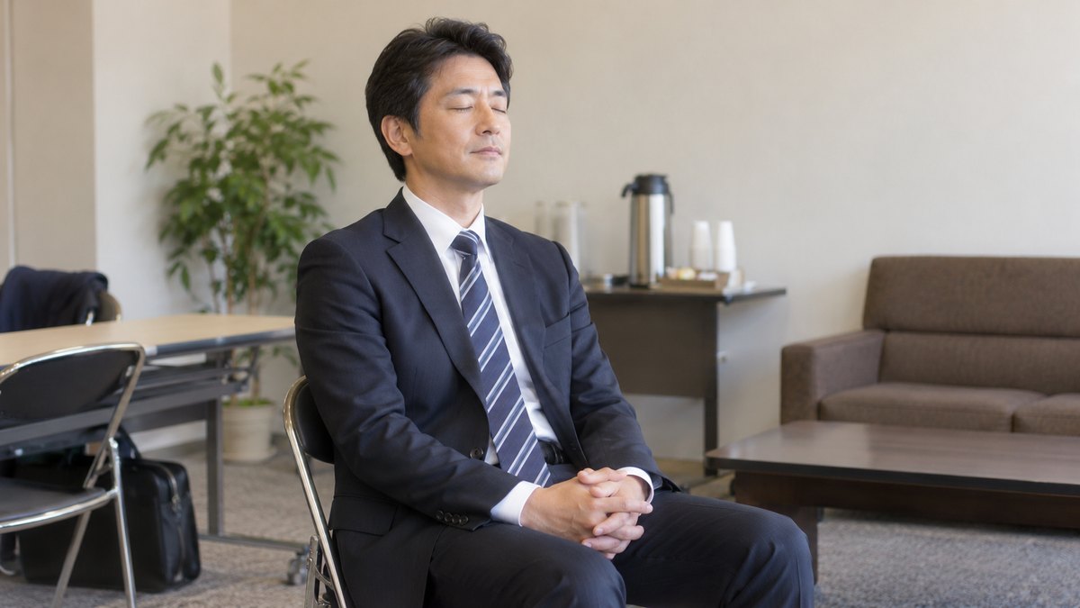 昼、市民ホールの控室で、パイプ椅子に座ったスーツ姿の40代の男性が、膝の上で手を軽く組み、目を閉じて呼吸を整えている。窓は写らない