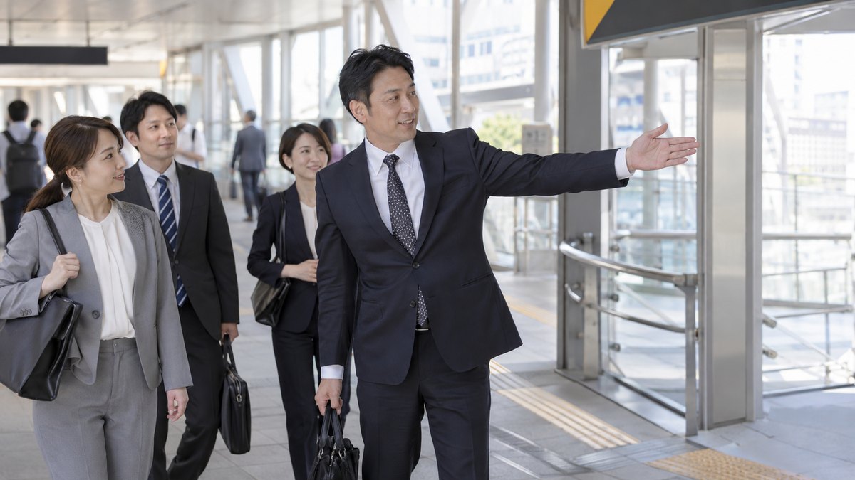 昼、駅の連絡通路で、スーツ姿の40代の男性が、通路の分かれ道に立って手で右の方向を示し、後ろに続く同僚たちを案内している。案内表示の文字は写らない
