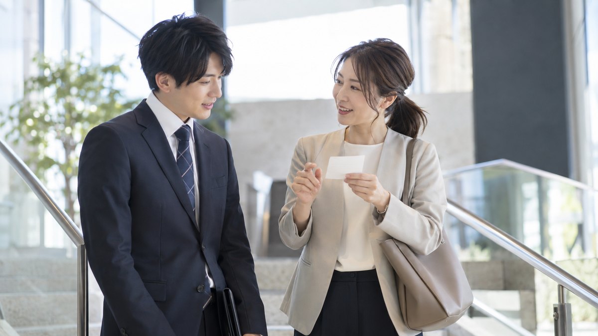 朝、会社のエントランスの階段で、30代の女性幹事が新しく来た20代の男性に小さなメモを見せながら、紹介の中身を確かめている。メモの文字は読めない