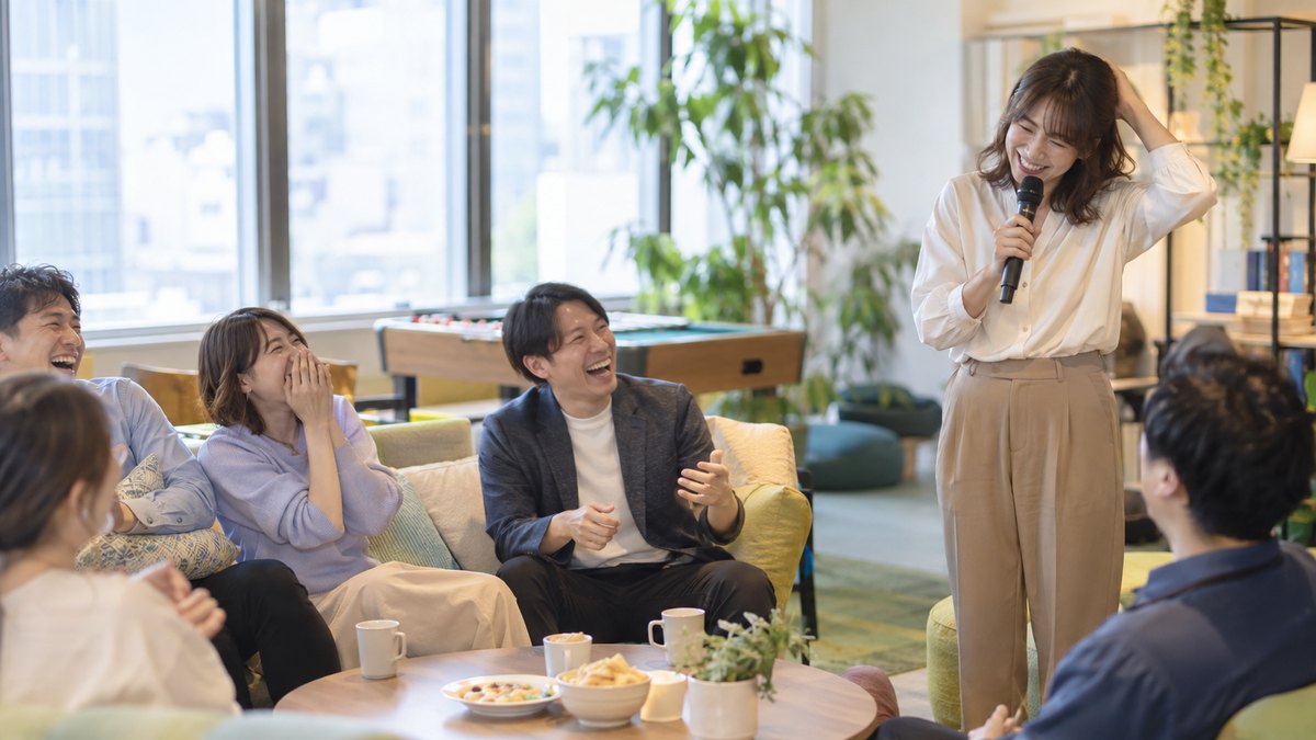 昼、会社のレクリエーションルームで、30代の女性司会者がマイクを持ったまま照れたように頭をかき、ソファに座った同僚たちが声を上げて笑っている