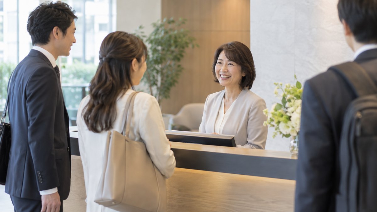 朝、会社の受付カウンターの内側で、50代の女性社員が、出社してきた社員たちにいつものように笑顔で声をかけている。看板や文字は写らない