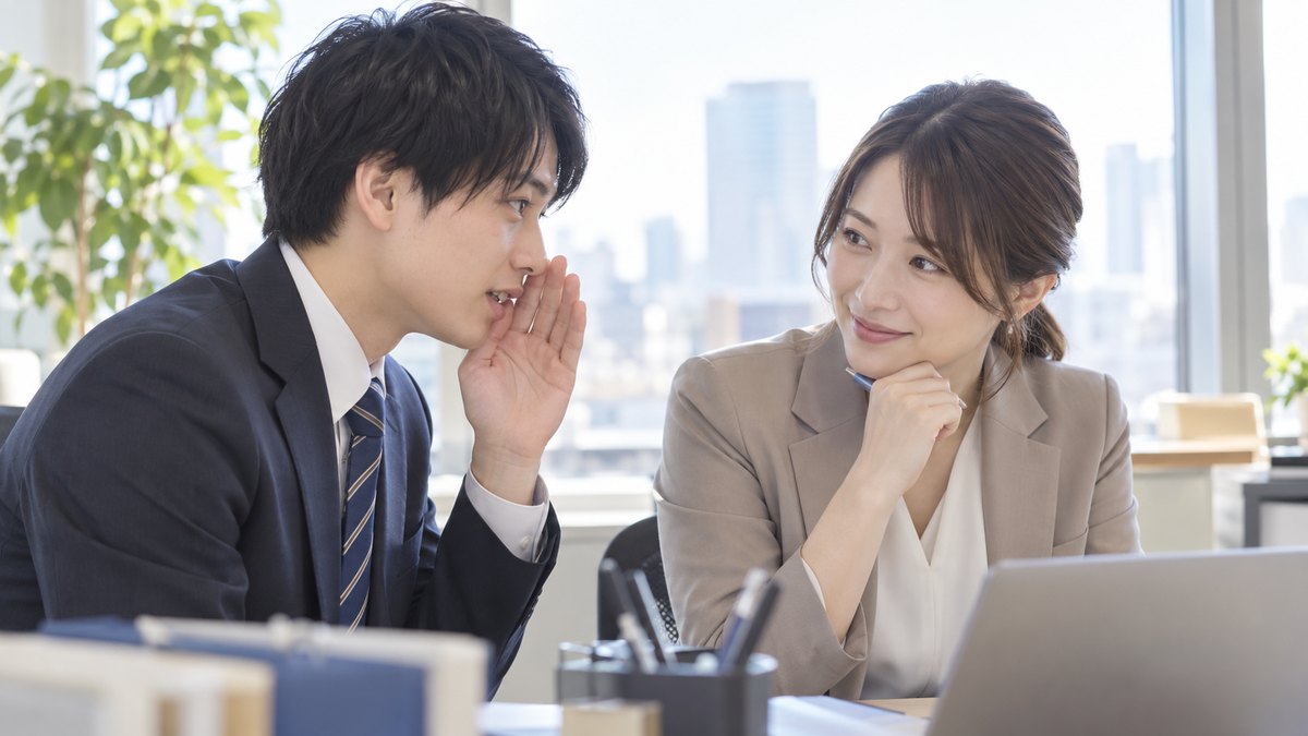 オフィスの窓際で、スーツ姿の20代の新人男性が、30代の先輩女性に忘年会で話す一言を小声で聞いてもらい、先輩がうなずいている