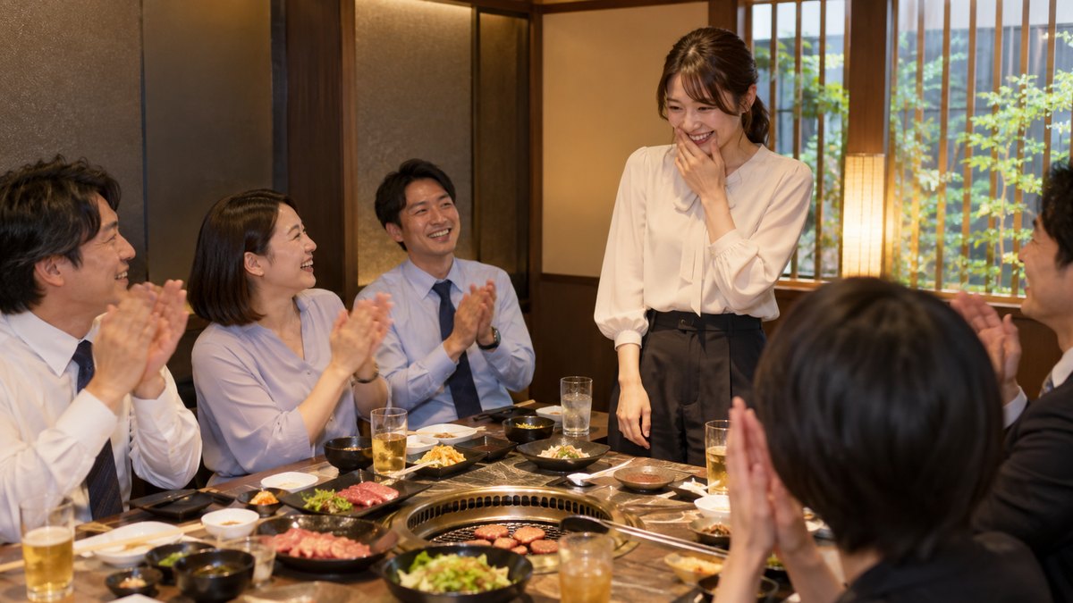 焼肉店の個室で、急に指名された30代の女性がトングを置いて少し照れたように立ち上がり、テーブルの同僚たちが拍手で迎えている