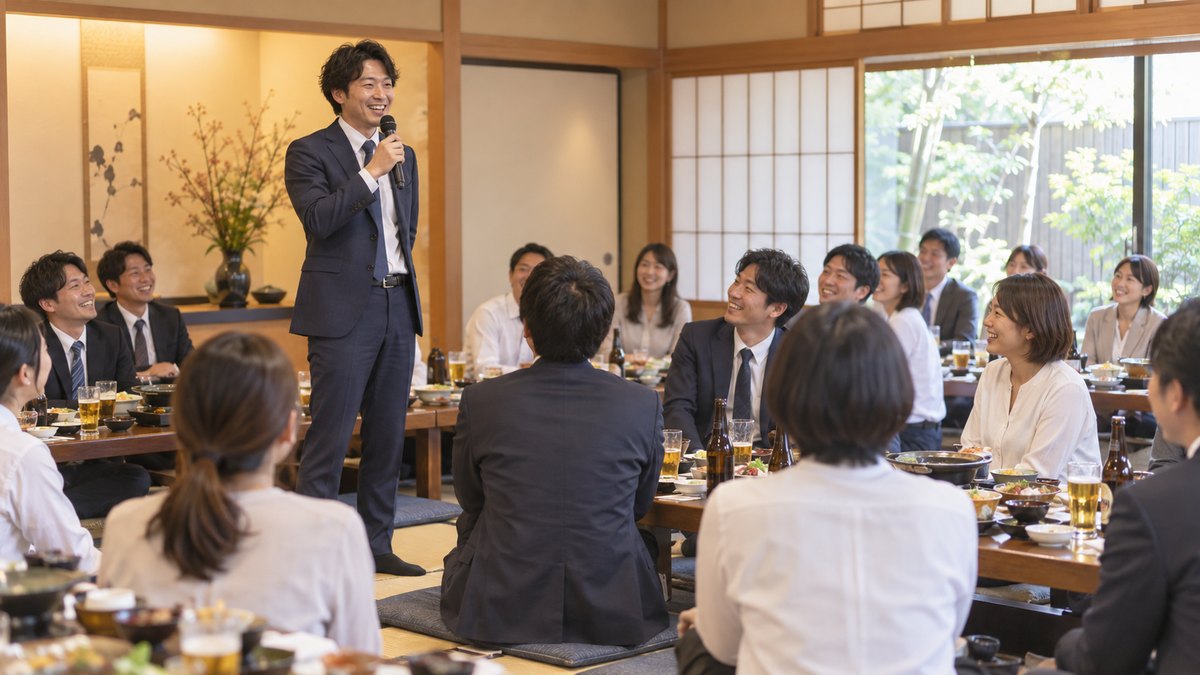 居酒屋の広い座敷の宴会で、30代の男性幹事がマイクを持って立ち、座卓を囲む20人ほどの社員に向かって笑顔で話し始めている