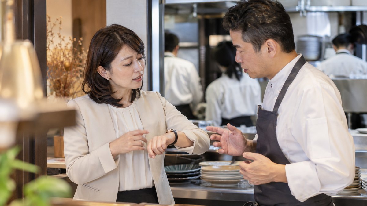 レストランの厨房の入口近くで、40代の女性幹事が腕時計を見ながら、店長と料理を出す時間を相談している